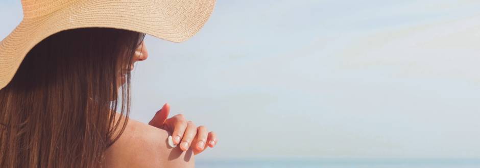 Lady applying sunscreen on her shoulder on beach wearing a big hat