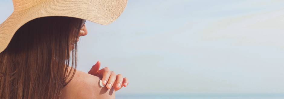 Lady applying sunscreen on her shoulder on beach wearing a big hat